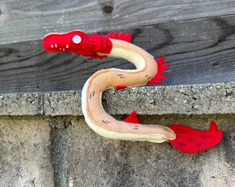 May include: A handmade plush toy in the shape of a sea serpent. The serpent features a red head and tail with a tan body. The toy is made of felt and has a whimsical design, perfect for a child's toy or room decor.