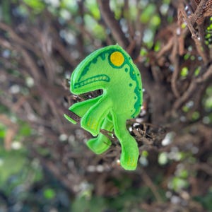 May include: A green felt monster with a yellow eye and green spots. The monster has a toothy grin and is posed with its arms and legs outstretched.