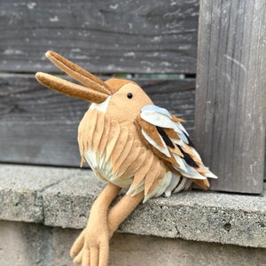 May include: A brown and beige felt bird with a long beak and outstretched wings. The bird is sitting on a gray concrete surface.