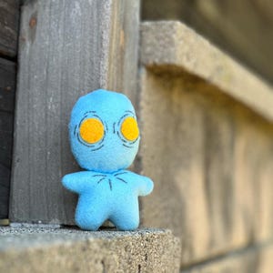 May include: A small blue plush toy with yellow eyes and a simple stitched design. The toy is standing on a gray surface.