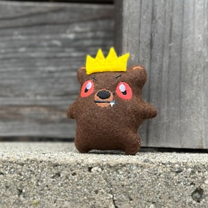 May include: A brown felt bear plush toy with a yellow crown. The bear has embroidered facial features, including red eyes, a black nose, and a small tooth. The toy is standing on a concrete surface.