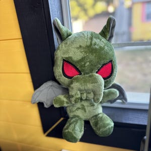 May include: Green plush toy with gray wings and red eyes. The toy is shaped like a creature with a large head and a small body.