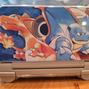 May include: A light gray handheld gaming console featuring a clear protective cover on the top lid. The cover displays a vibrant graphic illustration of the orange dragon-like creature Charizard and the blue turtle-like creature Blastoise.
