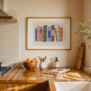 May include: A framed print featuring a collection of stylized books with colorful covers. The books have various patterns and colors, including stripes, diamonds, and floral designs. The print is displayed in a kitchen setting, above a wooden countertop.