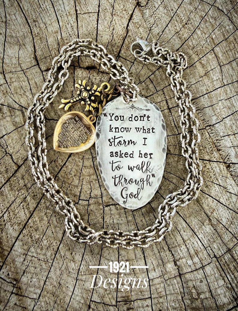 Pu&ograve; includere: Una collana color argento con un ciondolo ovale strutturato con l'iscrizione "You don't know what storm I asked her to walk through God." La collana include un ciondolo a forma di croce e cuore color oro. Lo sfondo &egrave; una superficie di legno rustico.