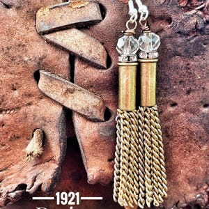 May include: A pair of gold tassel earrings with a clear crystal bead and rhinestones. The earrings are hanging from a brown leather background. The text "1921 Designs" is visible at the bottom of the image.