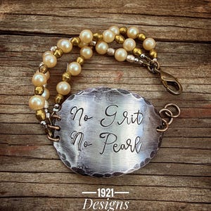 May include: A handmade bracelet featuring a silver oval pendant with the engraved text "No Grit No Pearl." The bracelet has strands of cream-colored pearls and gold-colored beads. The clasp is a bronze color. The bottom of the image has the text "1921 Designs."