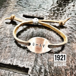 May include: A brown leather bracelet with a silver bead and a metal oval charm that says "G>AV". The bracelet is on a wooden surface. 1921 Designs.