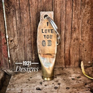 May include: A brass keychain with a bullet-shaped charm engraved with "I Love You GB". The keychain is attached to a silver key ring. The charm is on a wooden surface with the text "1921 Designs" below it.