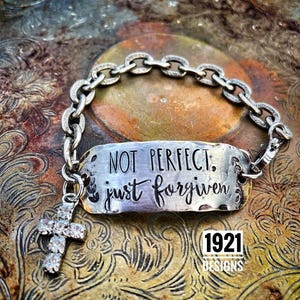 May include: Silver bracelet with a charm that reads "Not perfect, just forgiven." The charm is attached to a silver chain with a small cross charm with rhinestones. The bracelet is on a textured gold surface. 1921 Designs is printed below the bracelet.