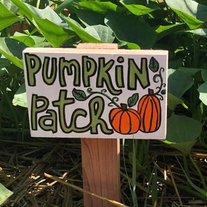 May include: A wooden sign with the words "Pumpkin Patch" written in green and brown lettering. Two orange pumpkins are drawn on the sign. The sign is placed in a garden bed with green leaves and straw mulch.