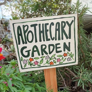 May include: A white wooden sign with green lettering that reads "Apothecary Garden". The sign is decorated with small pink and orange flowers and green leaves. The sign is attached to a wooden stake.