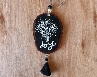May include: A black wooden round ornament with a white floral design and the word "joy" painted on it. The ornament has a black tassel and a twine loop for hanging.