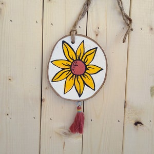 May include: A wooden round ornament with a painted yellow sunflower with a red center. The ornament has a brown twine hanger and a red tassel.