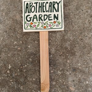 May include: A white ceramic garden marker with a wooden stake. The marker has green lettering that says "Apothecary Garden" and is decorated with small pink and orange flowers.