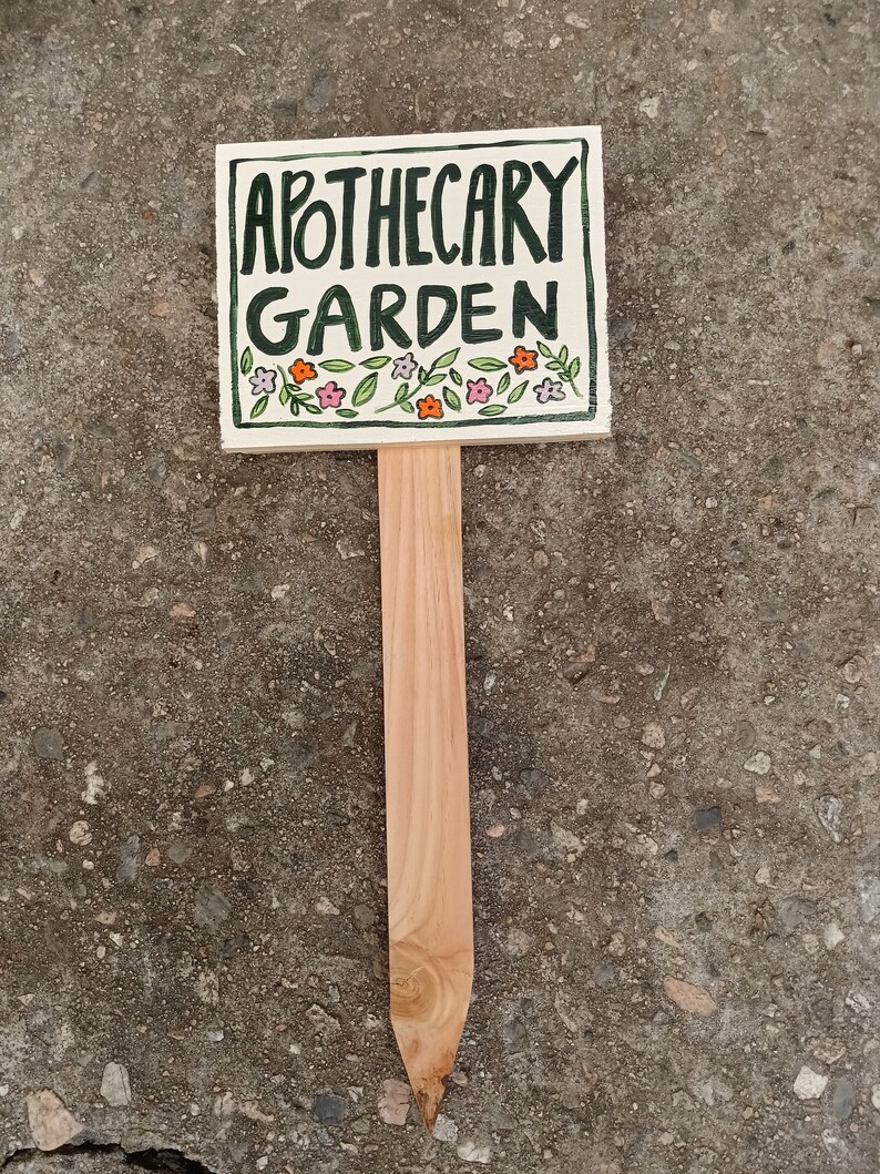 May include: A white ceramic garden marker with a wooden stake. The marker has green lettering that says "Apothecary Garden" and is decorated with small pink and orange flowers.