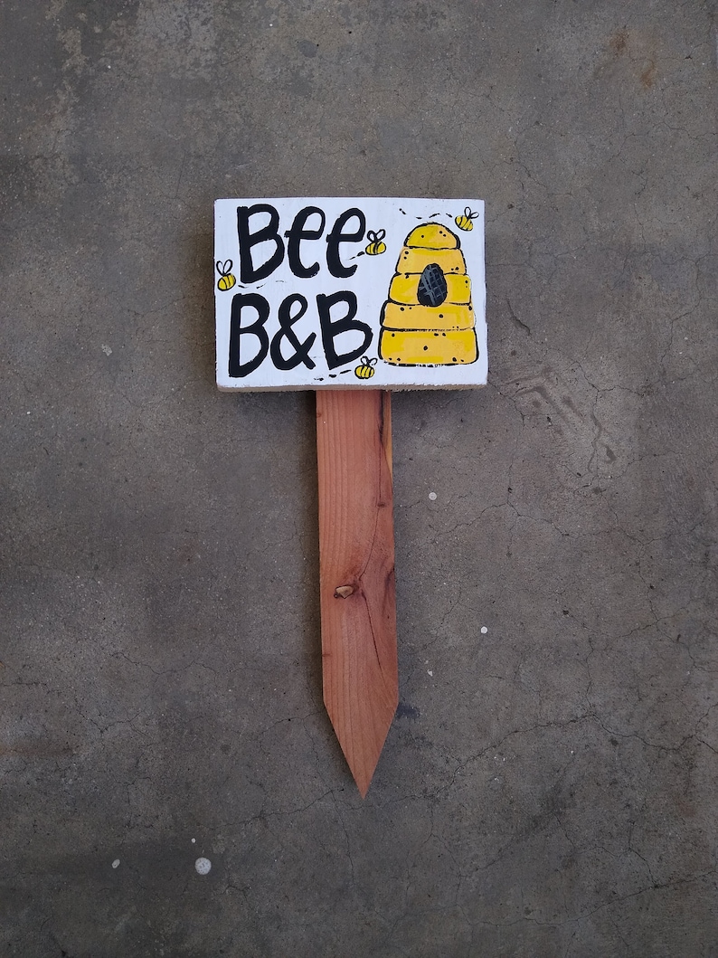 May include: A wooden sign with the words "Bee B&B" in black lettering, along with a yellow beehive illustration and small bee drawings. The sign is mounted on a wooden stake, perfect for a garden or yard.