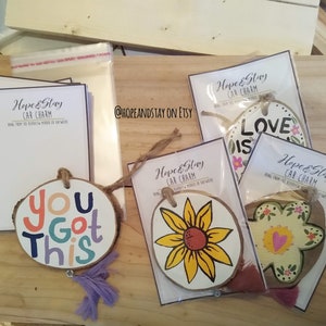 May include: Four wooden car charms with different designs. The charms are painted with bright colors and have a rustic look. The designs include a sunflower, a flower with a heart, a flower with the words "Love is", and the words "You Got This".