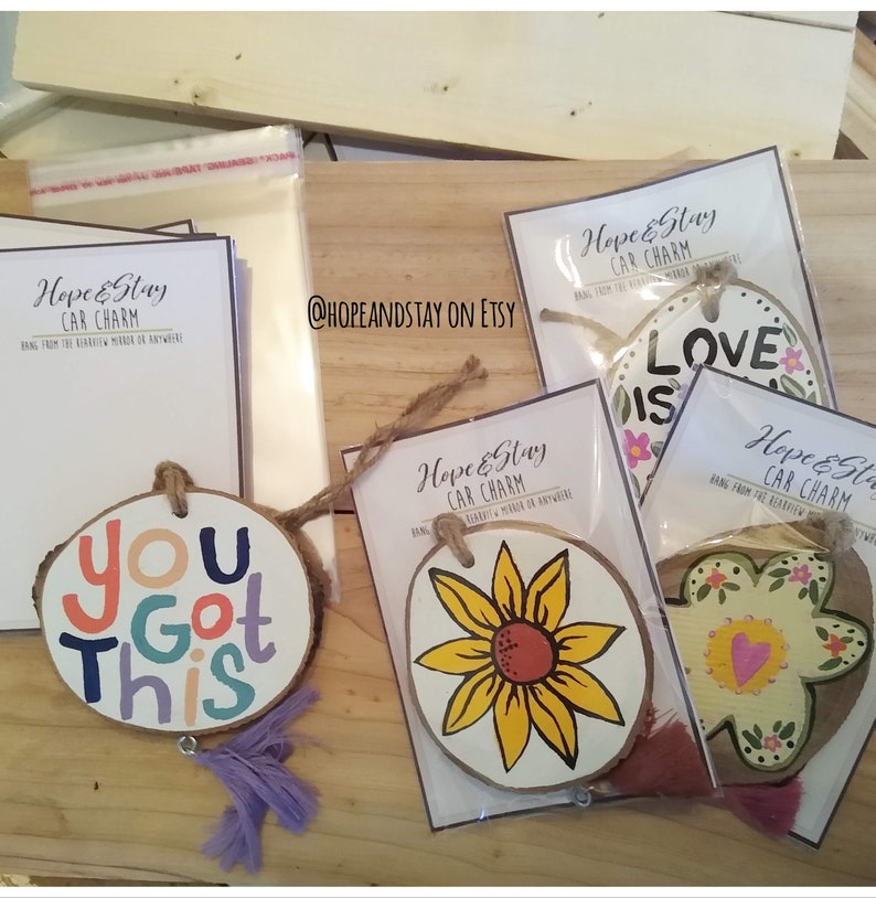 May include: Four wooden car charms with different designs. The charms are painted with bright colors and have a rustic look. The designs include a sunflower, a flower with a heart, a flower with the words "Love is", and the words "You Got This".