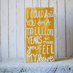 May include: A yellow wooden sign with white lettering that reads "I could hold you for a million years to make you feel my love".