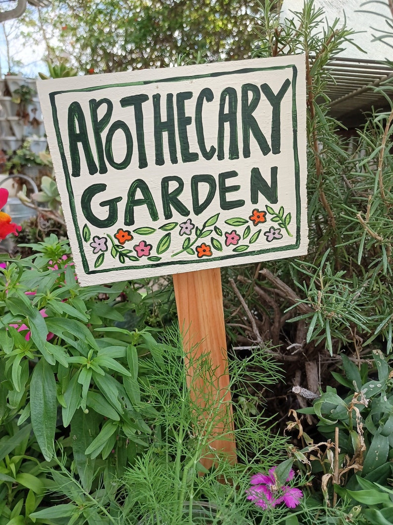May include: A white wooden sign with green lettering that reads "Apothecary Garden". The sign is decorated with small pink and orange flowers and green leaves. The sign is attached to a wooden stake.
