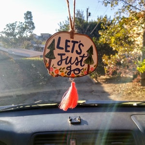May include: A wooden car air freshener with the text "Let's Just Go" painted on it. The wood is painted to look like a tree trunk with green trees and pink flowers painted around the text. The air freshener has a pink tassel hanging from it.