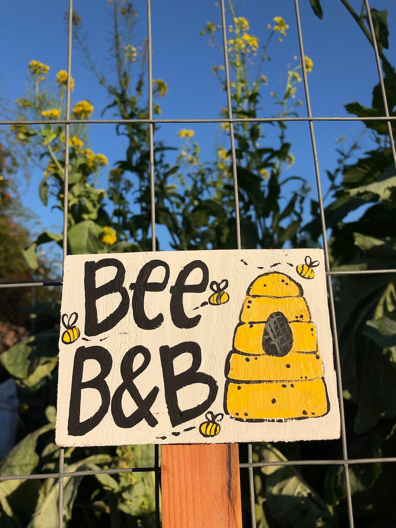 May include: A hand-painted wooden sign with the words "Bee B&B" in black lettering, featuring a yellow beehive and cartoon bees. The sign is mounted on a wooden post against a backdrop of a wire fence, yellow flowers, and a blue sky.