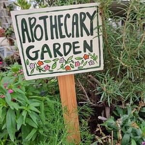 May include: A white wooden sign with green lettering that reads "Apothecary Garden". The sign is decorated with small pink, orange, and green flowers and leaves. The sign is attached to a wooden stake and is placed in a garden.