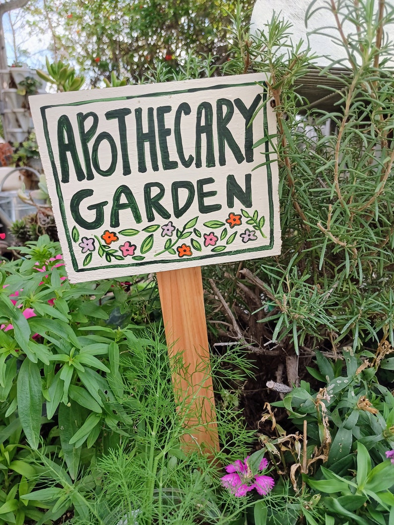 May include: A white wooden sign with green lettering that reads "Apothecary Garden". The sign is decorated with small pink, orange, and green flowers and leaves. The sign is attached to a wooden stake and is placed in a garden.