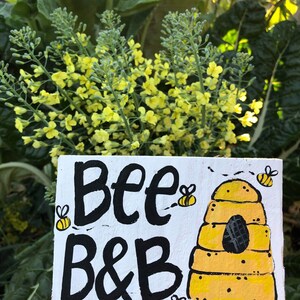 May include: A white wooden sign with the words "Bee B&B" in black lettering, accompanied by a yellow and black beehive illustration and three cartoon bees. The sign is set against a backdrop of green foliage and yellow flowers.