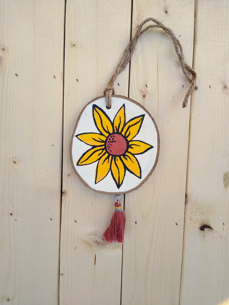 May include: A wooden round ornament with a painted yellow sunflower with a red center. The ornament has a brown twine hanger and a red tassel.