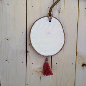 May include: A round wooden slice painted white with a red tassel hanging from it. The slice is attached to a piece of twine.