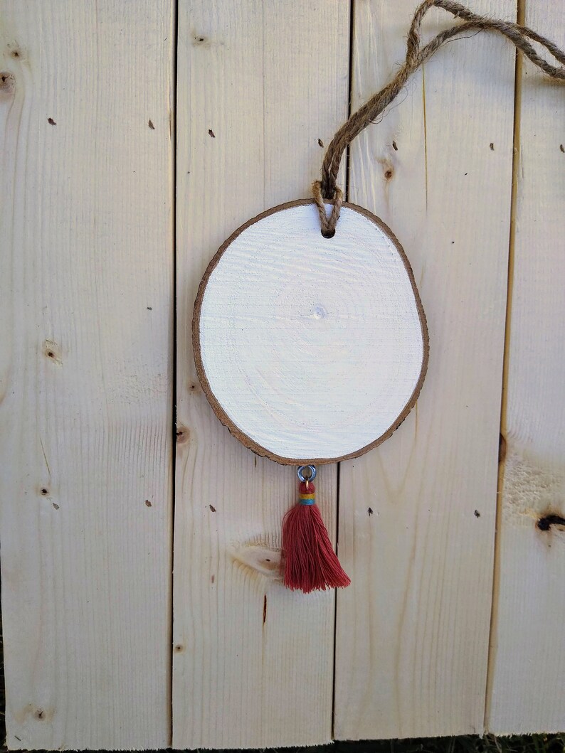 May include: A round wooden slice painted white with a red tassel hanging from it. The slice is attached to a piece of twine.