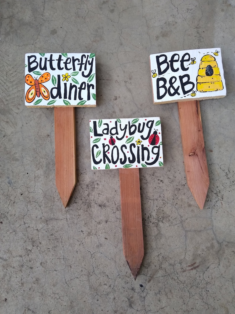 May include: Three wooden garden signs with hand-painted designs. One sign reads "Butterfly diner" with a butterfly illustration. Another says "Bee B&B" with a beehive. The third sign says "Ladybug Crossing" with ladybug art.