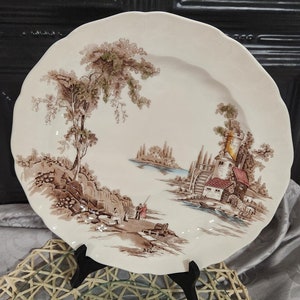 May include: A vintage oval-shaped ceramic plate with a brown and white landscape scene. The plate features a brown tree, a small cottage, and a river. The plate is on a stand and a glass plate.