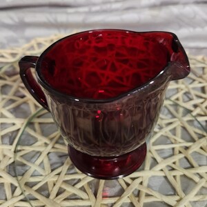 May include: A red glass creamer with a textured design. The creamer has a handle and a round base.