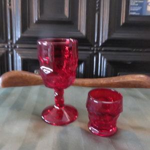 May include: Two red glass goblets, one tall and one short, with a textured, faceted design. The goblets are on a green tablecloth.