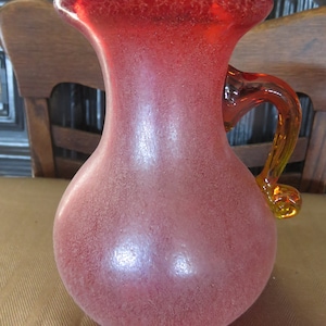 May include: A frosted glass pitcher with a textured surface. The pitcher is a deep red color with a yellow and orange handle.