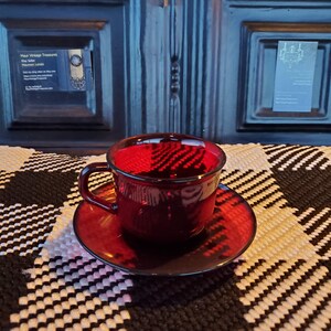 May include: A red glass teacup and saucer set. The cup and saucer are both a deep red color and have a glossy finish.