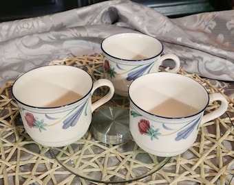 May include: Three white ceramic teacups with blue trim and floral designs. The cups are on a woven placemat with a glass surface underneath.