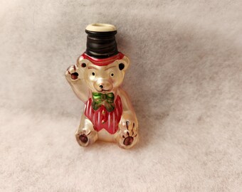 May include: A vintage glass Christmas ornament shaped like a bear. The bear wears a black top hat, a red vest with white stripes, and a green bow tie. The bear is waving with its right paw.