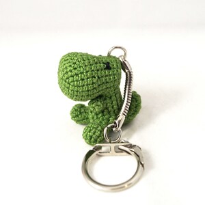 May include: Green crocheted dinosaur keychain with a silver metal ring and chain.