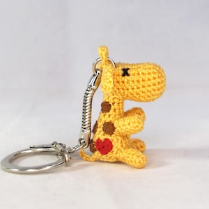 May include: A crocheted yellow giraffe keychain with brown spots and a red heart. The giraffe has a silver key ring and a spring-loaded connector. The keychain is handmade and has a cute, whimsical design.