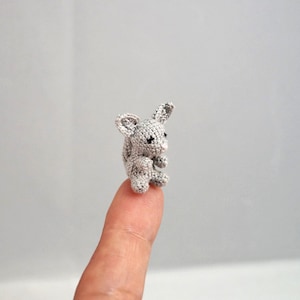 May include: A small grey crocheted animal with large ears and black eyes. The animal is sitting on a finger.