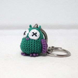 May include: Green crocheted owl keychain with purple accents and white eyes. The owl has a stitched X on each eye. The keychain is attached to a silver metal ring.