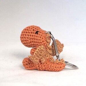 May include: A small crocheted orange dragon keychain with a silver spring clasp. The dragon has tan wings and a black embroidered eye.