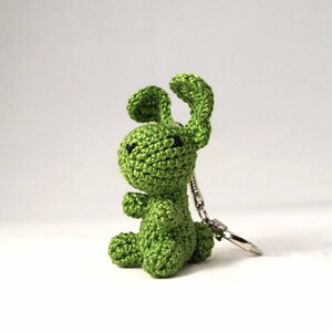 May include: A green crocheted bunny keychain. The bunny is sitting up with its ears perked up. It has black eyes and a silver keychain attached.