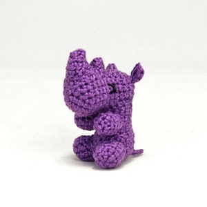 May include: A small purple crocheted rhinoceros toy. The toy is sitting on a white surface.