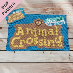 Może przedstawiać: Koc wykonany na szydełku z logo Animal Crossing z napisami "Animal Crossing" i "New Horizons" w żółtej i brązowej włóczce na niebieskim tle.