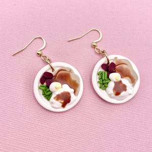 May include: A pair of dangle earrings featuring miniature food designs. Each earring has a white plate with a brown meat, red beans, green chives, and a dollop of white sauce with brown gravy. The earrings are suspended from gold-colored hooks.